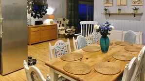Uma área de refeições com mesa de madeira, jogos americanos trançados, cadeiras brancas, um vaso azul com flores e uma cozinha com geladeira e armários de madeira.