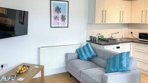 Uma sala de estar moderna com um sofá cinzento, almofadas azuis, uma mesa de centro de madeira com frutas e uma revista, uma TV na parede e uma área de cozinha com armários brancos e um micro-ondas.