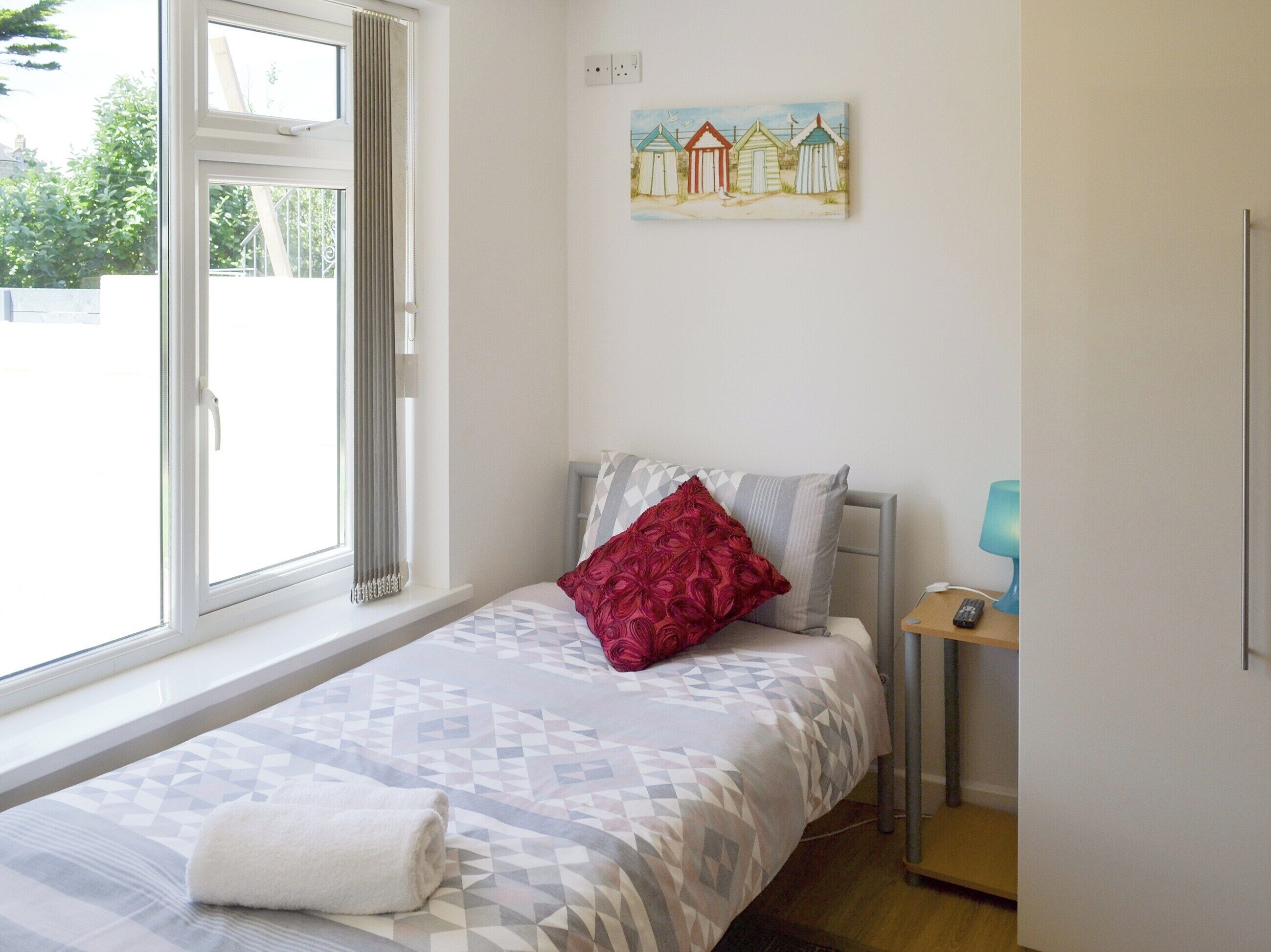 Een slaapkamer met een bed, een raam met gordijnen, een nachtkastje met een lamp, en een schilderij aan de muur.