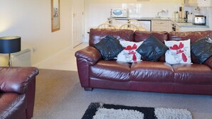 A living room with a leather sofa, a coffee table, and a lamp. There is a kitchen in the background with a microwave, toaster, and cabinets. - 3 bedroom accommodation in Whitby (Whitby)