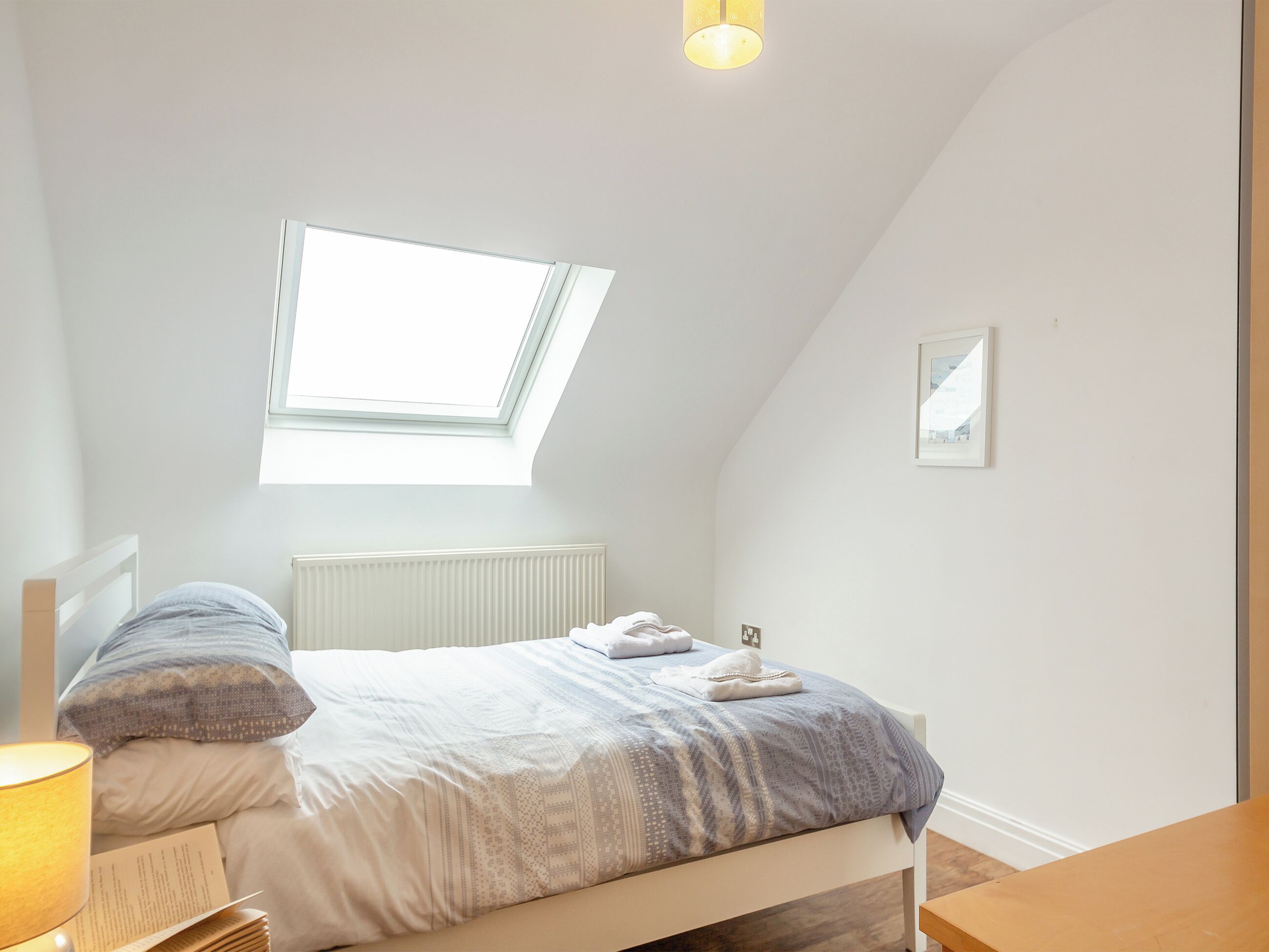 A bedroom with a skylight, a bed with a patterned bedspread, a bedside lamp, and a framed picture on the wall.