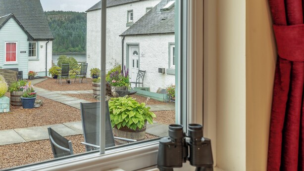 A window view of a courtyard with a white building, a wooden bench, and potted plants. - Barn Owl Cottage - 27941 (Stirling)