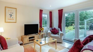 A living room with a television, a coffee table, a sofa, and a chair. There is a window with curtains, and a door leading to a balcony. - Barn Owl Cottage - 27941 (Stirling)