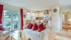 A modern living room with a white sofa, red cushions, and a coffee table. A kitchen with white cabinets and a dining area is visible in the background. - Barn Owl Cottage - 27941 (Stirling)
