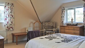 Une chambre avec un plafond en pente, un lit recouvert d’une couvre-lit à motifs floraux, une commode, une chaise et une fenêtre avec des rideaux.