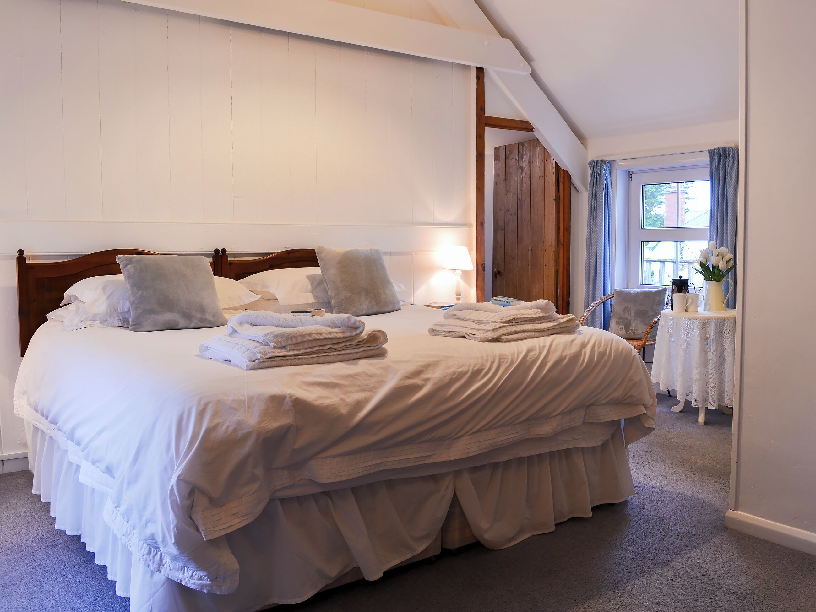 Een netjes opgemaakt bed met kussens en opgevouwen handdoeken, een houten hoofdbord, een nachtkastje met een lamp en een raam met gordijnen.