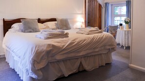 Ein ordentlich bezogenes Bett mit Kissen und gefalteten Handtüchern, ein Holz-Kopfteil, ein Nachttisch mit Lampe und ein Fenster mit Vorhängen.