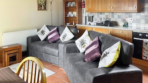 Ein Wohnzimmer mit einer grauen Sofagarnitur, einem Holztisch und einem Sessel. Im Hintergrund ist eine Küche mit Holzschränken und einer gefliesten Spritzschutzleiste zu sehen.