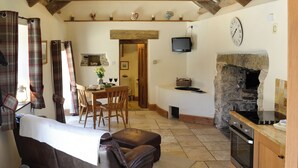 A cozy living room with a sofa, a dining area with a table and chairs, a kitchen with a stone wall and an oven, and a television mounted on the wall. - 1 bedroom accommodation in Mevagissey (Mevagissey)