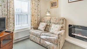 A cozy living room with a sofa, a television, a fireplace, and a window with curtains. - 2 bedroom accommodation in Pembrey, near Burry Port (Pembrey, near Burry Port)