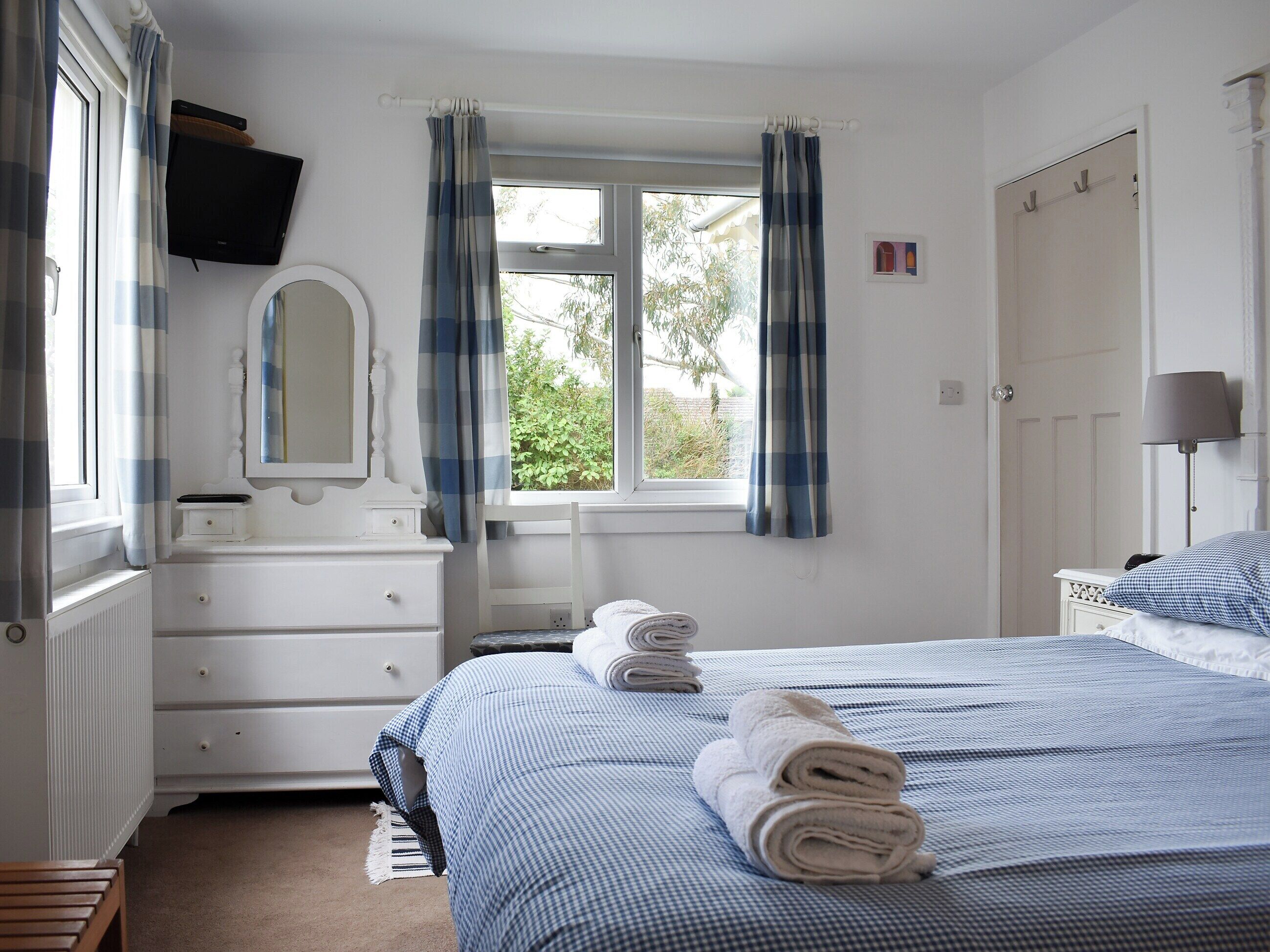 Une chambre à coucher comprenant un lit, une commode, un miroir, une fenêtre avec des rideaux, une lampe de chevet et une vue sur la verdure à l’extérieur.