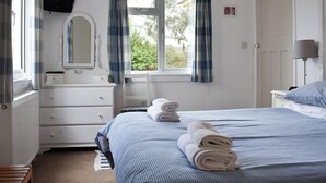 Een slaapkamer met een bed, een dressoir, een spiegel, een raam met gordijnen, een bedlampje en uitzicht op groen buiten.