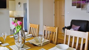 Un coin repas avec une table en bois dressée pour quatre personnes, un vase de fleurs et, au fond, une cuisine moderne.
