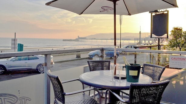 Une table entourée de chaises et protégée par un parasol, donnant sur une plage où l’on aperçoit des voitures et, au loin, une jetée.