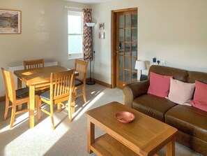 A living room with a wooden table, chairs, a brown leather sofa, a floor lamp, and a window with curtains. - An attractive luxurious ground floor apartment in the centre of Keswick. (Keswick)