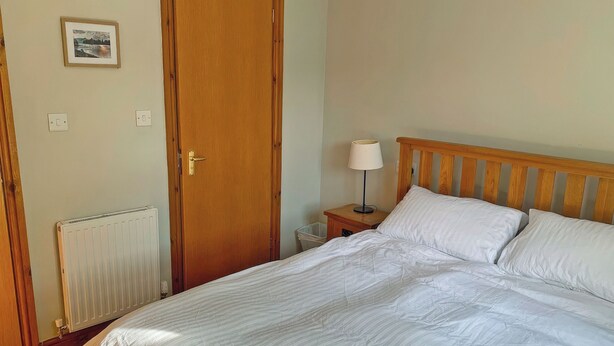 A bedroom with a wooden bed, a nightstand with a lamp, a radiator, a door, and a framed picture on the wall. - An attractive luxurious ground floor apartment in the centre of Keswick. (Keswick)