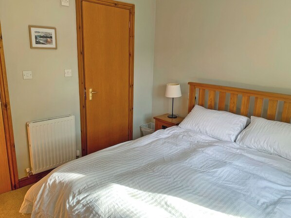 A bedroom with a wooden bed, a nightstand with a lamp, a radiator, a door, and a framed picture on the wall. - An attractive luxurious ground floor apartment in the centre of Keswick. (Keswick)
