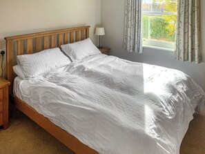 A bedroom with a wooden bed frame, white bedding, a bedside lamp, and a window with curtains. - An attractive luxurious ground floor apartment in the centre of Keswick. (Keswick)