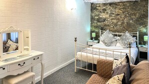 Un dormitorio con pared de ladrillo blanco, pared de acento de piedra, cama blanca con múltiples almohadas, mesita de noche con espejo y una pequeña mesita con lámpara.