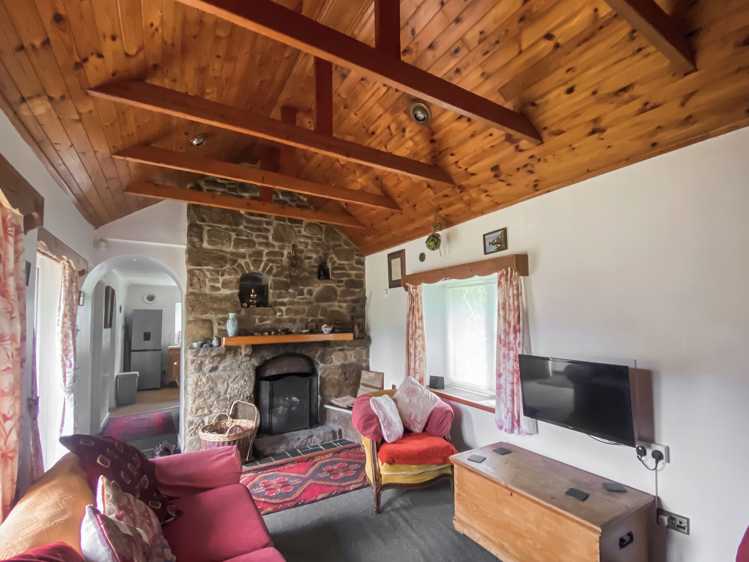 Un salon chaleureux avec une cheminée en pierre, des poutres apparentes en bois au plafond, un confortable canapé rouge, un téléviseur et un mur en pierre.