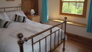 A bedroom with a wooden bed frame, a nightstand with a lamp, a window with a view of greenery, and a wooden wardrobe. - Gateside Farmhouse Sedbergh (Sedbergh)