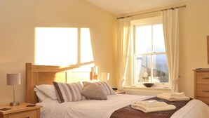 A neatly made bed with a brown and white striped pillow, a wooden nightstand with a lamp, and a window with curtains. - 2 bedroom accommodation in Thornthwaite (Thornthwaite)