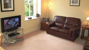 A living room with a brown leather sofa, a television on a glass stand, a window with a view of trees, and a lamp on a side table. - 2 bedroom accommodation in Thornthwaite (Thornthwaite)