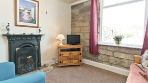 A living room with a fireplace, a television on a wooden stand, a stone wall, and a window with curtains. - 3 bedroom accommodation in Staithes, near Whitby (Staithes, near Whitby)