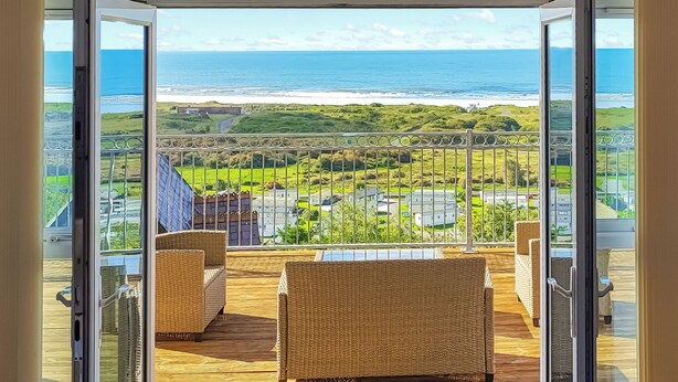 A balcony with wicker furniture, a glass door, and a view of the ocean and greenery. - Pendine Beach Apartment (Pendine, near Amroth)