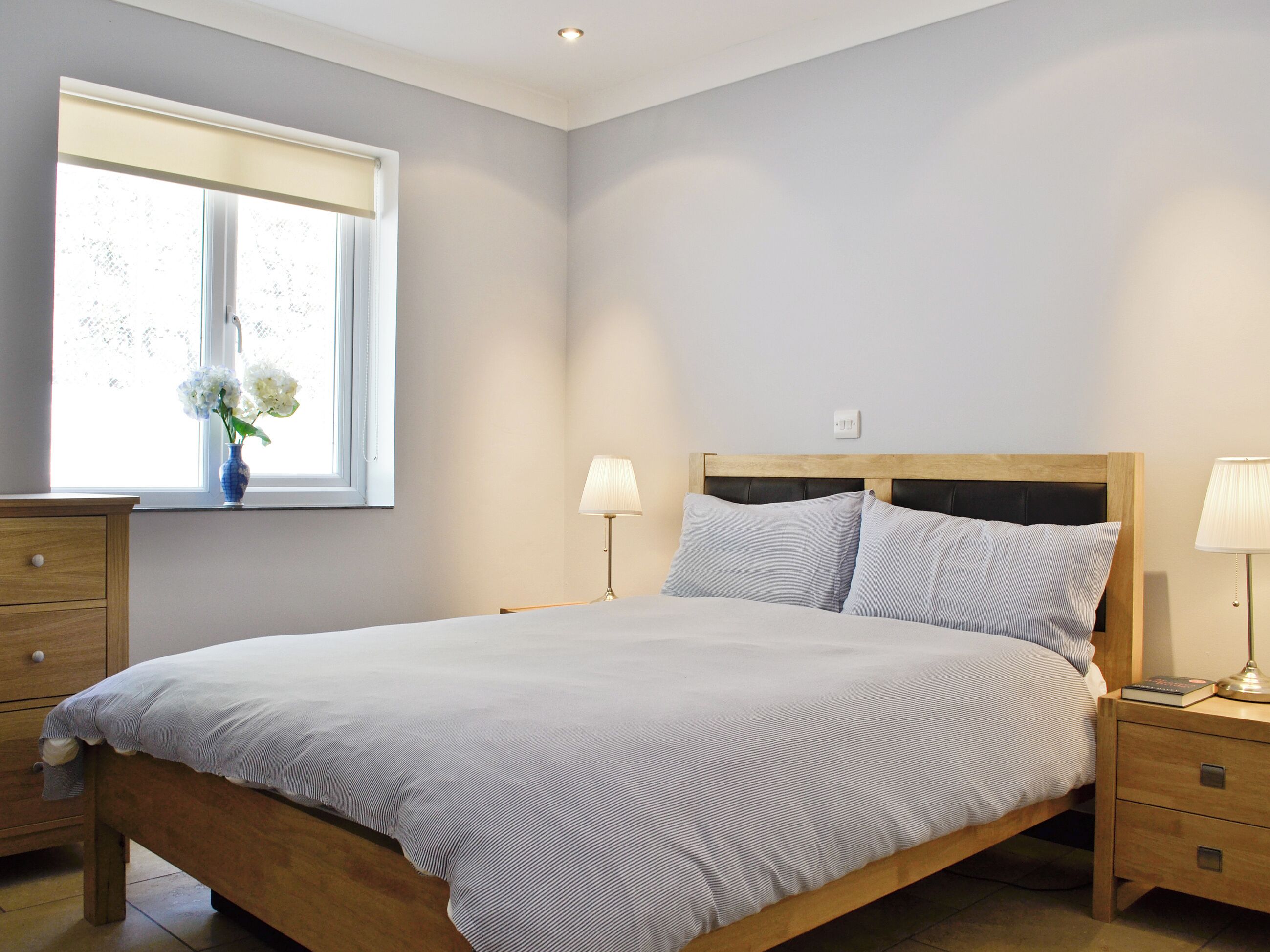 Une chambre à coucher avec un lit, des tables de chevet, une fenêtre et un vase rempli de fleurs.