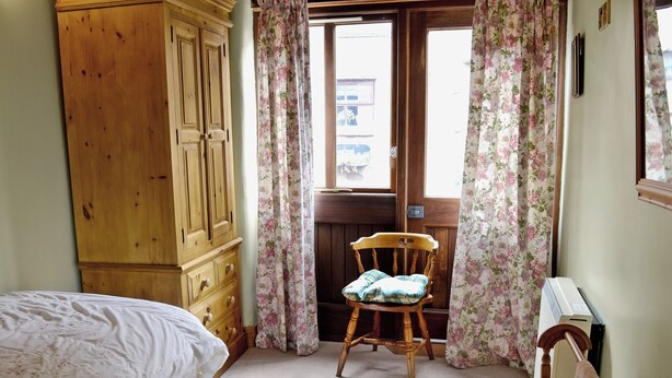 Um quarto com guarda-roupa de madeira, uma cama com colcha branca, uma cadeira de madeira com almofada e uma janela com cortinas florais.
