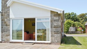Ein kleines, eingeschossiges Haus mit Natursteinaußenwand und weißer Fassade. Es hat eine Schiebetür aus Glas, die zu einem Wohnbereich mit Sofa und Fernseher führt.