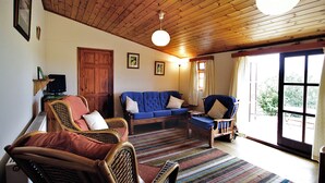 Ein gemütliches Wohnzimmer mit Holzbalkendecke, einem blauen Sofa, Korbstühlen und einem großen Fenster mit Vorhängen.