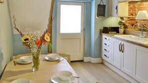 Ein Essbereich mit Tisch für vier Personen, einer Blumenvase und einer Küche mit weißen Schränken und einer Ziegelrückwand.