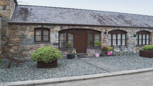 A stone building with a sloped roof, a wooden door, and multiple windows. There are potted plants and a metal bench in front. - 2 bedroom accommodation in Caernarfon (Caernarfon)