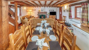 Un coin repas est aménagé avec une longue table en bois, dressée pour six personnes, et entourée de chaises en bois. On aperçoit une cheminée au fond, et un téléviseur est fixé au mur.