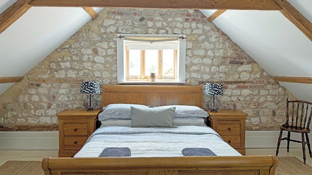 A bedroom with a sloped ceiling, stone wall, wooden bed frame, two bedside tables, a chair, and a window with a view. - 6 bedroom accommodation in Oxborough (Oxborough)