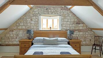 A bedroom with a sloped ceiling, stone wall, wooden bed frame, two bedside tables, a chair, and a window with a view.