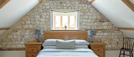 A bedroom with a sloped ceiling, stone wall, wooden bed frame, two bedside tables, a chair, and a window with a view.