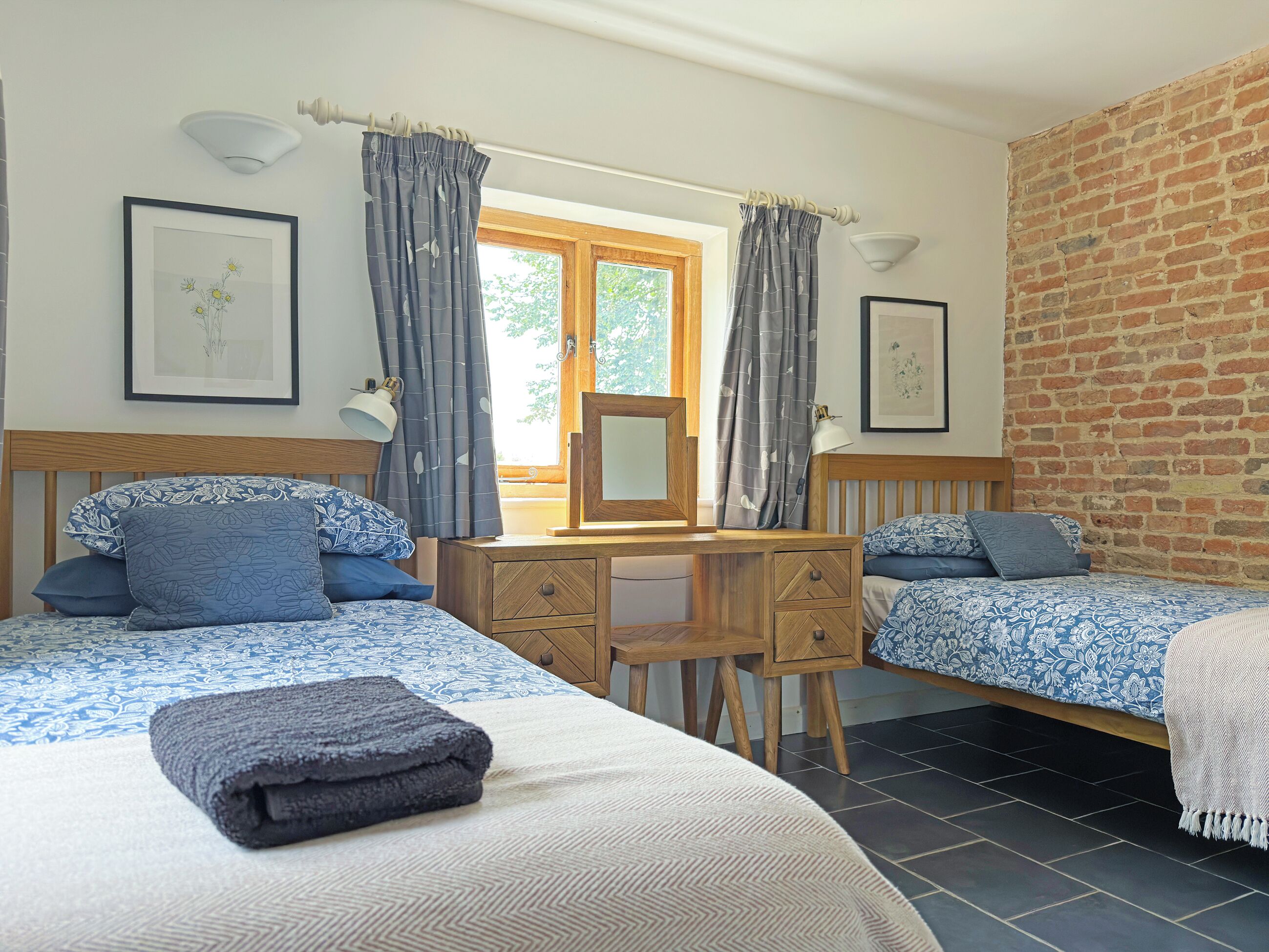 A bedroom with two beds, a wooden desk, a chair, a mirror, and a window with curtains.