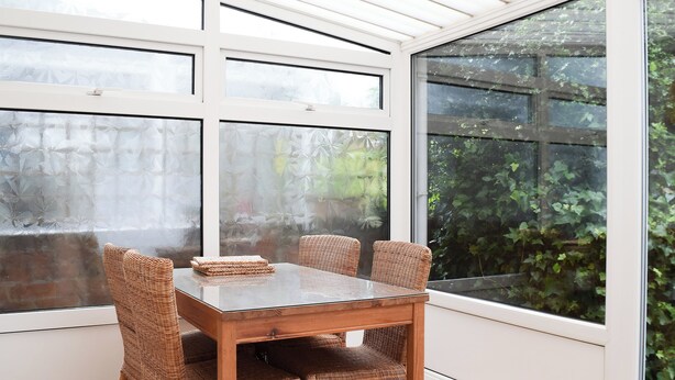 Een eetruimte met een glazen tafel, rieten stoelen, grote ramen en uitzicht op groen buiten.