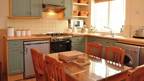 Een keuken met blauwgroene kastjes, een fornuis, een spoelbak en een vaatwasser. Op de planken staan rieten manden en er staat een houten tafel met stoelen.
