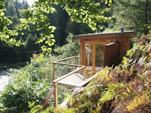 A wooden cabin with a glass door, surrounded by dense foliage and a river. - 4 bedroom accommodation in Seldom Seen near Thornthwaite (Seldom Seen near Thornthwaite)