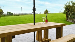 Une table de pique-nique en bois, avec une bouteille et un verre dessus, placée sous un grand parasol, sur une terrasse pavée, avec un champ d’herbe en arrière-plan.