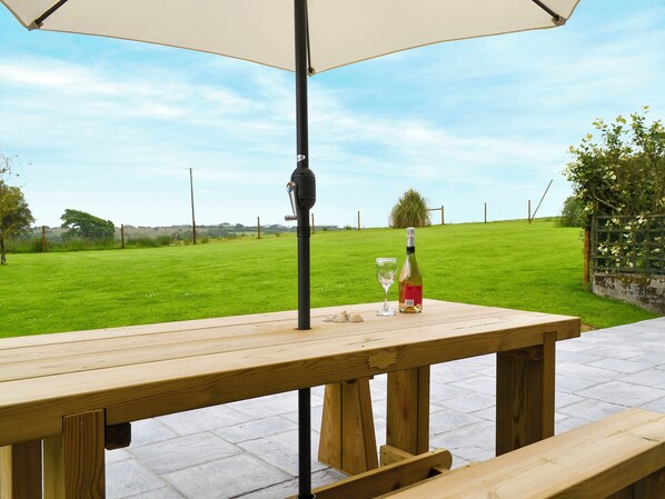 Uma mesa de piquenique de madeira com uma garrafa e um copo, sob um grande guarda-sol, num pátio pavimentado com um campo gramado ao fundo.