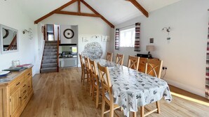 Un espace repas avec une longue table, des chaises en bois et un mur en pierre. Un escalier mène à une autre pièce, et on aperçoit un coin cuisine au fond.