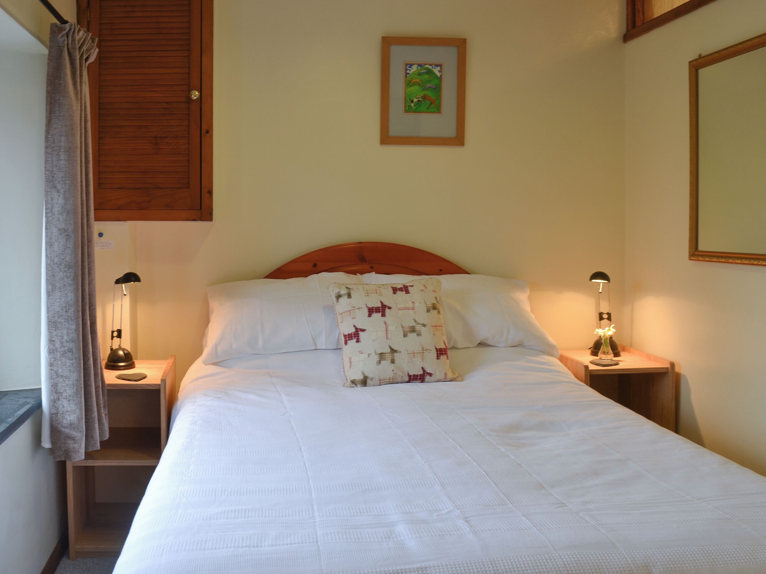 A bedroom with a wooden headboard, a bed with white bedding, two bedside lamps, a nightstand, a framed picture on the wall, and a window with curtains.