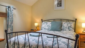 Een slaapkamer met een bed, nachtkastje, lamp, een lijst met een afbeelding aan de muur en een raam met gordijn.