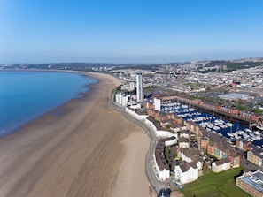 An aerial view of a coastal town with a long sandy beach, a marina with numerous boats, and a mix of residential and commercial buildings.
