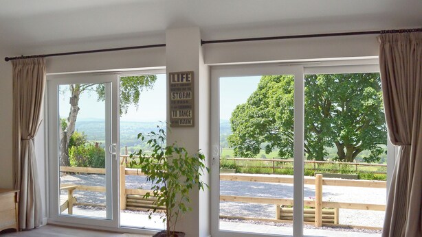 Ein Zimmer mit großen Schiebeglasfenstern, Holzboden, einer Topfpflanze und Blick auf eine Landschaft mit Bäumen und einem Zaun.